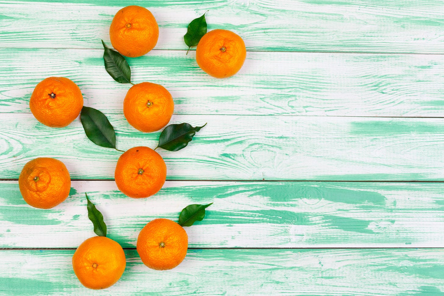 Eight oranges with green leaves are arranged on a light green wooden table.