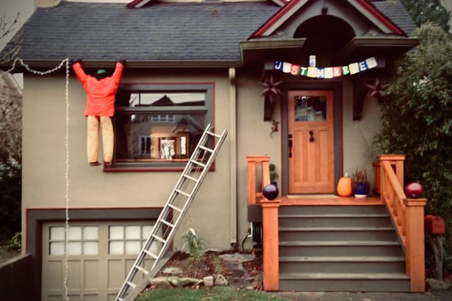 A man appears to be hanging from the gutter of his house with his ladder fallen off to the side.