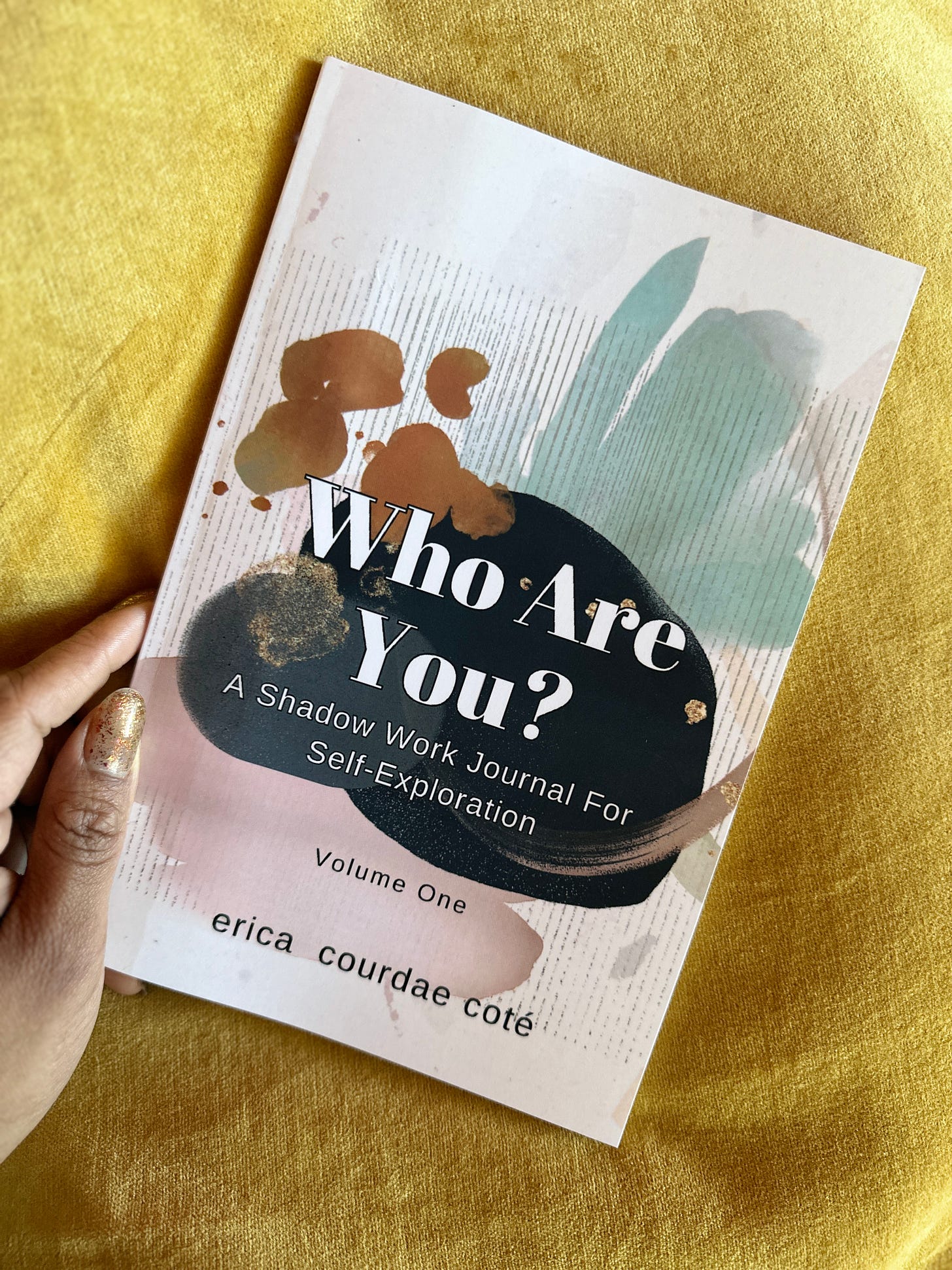 A Black woman's hand holding a copy of the book Who Are You? A Shadow Work Journal for Self-Exploration on a gold velvet pillow