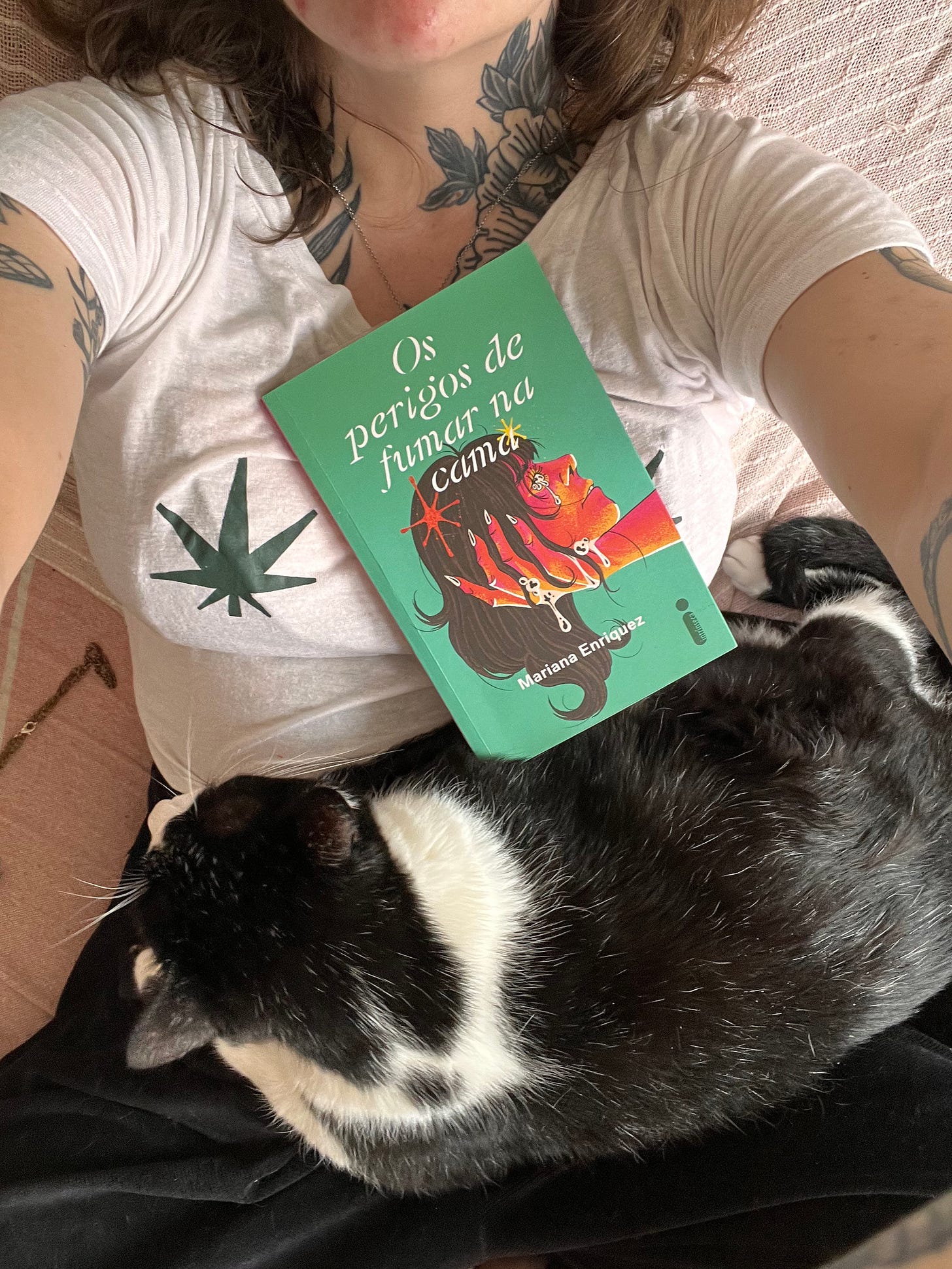 Foto do livro Os perigos de fumar na cama, tirada de cima, o livro está em cima de mim e tem um gato deitado na minha barriga próximo dele