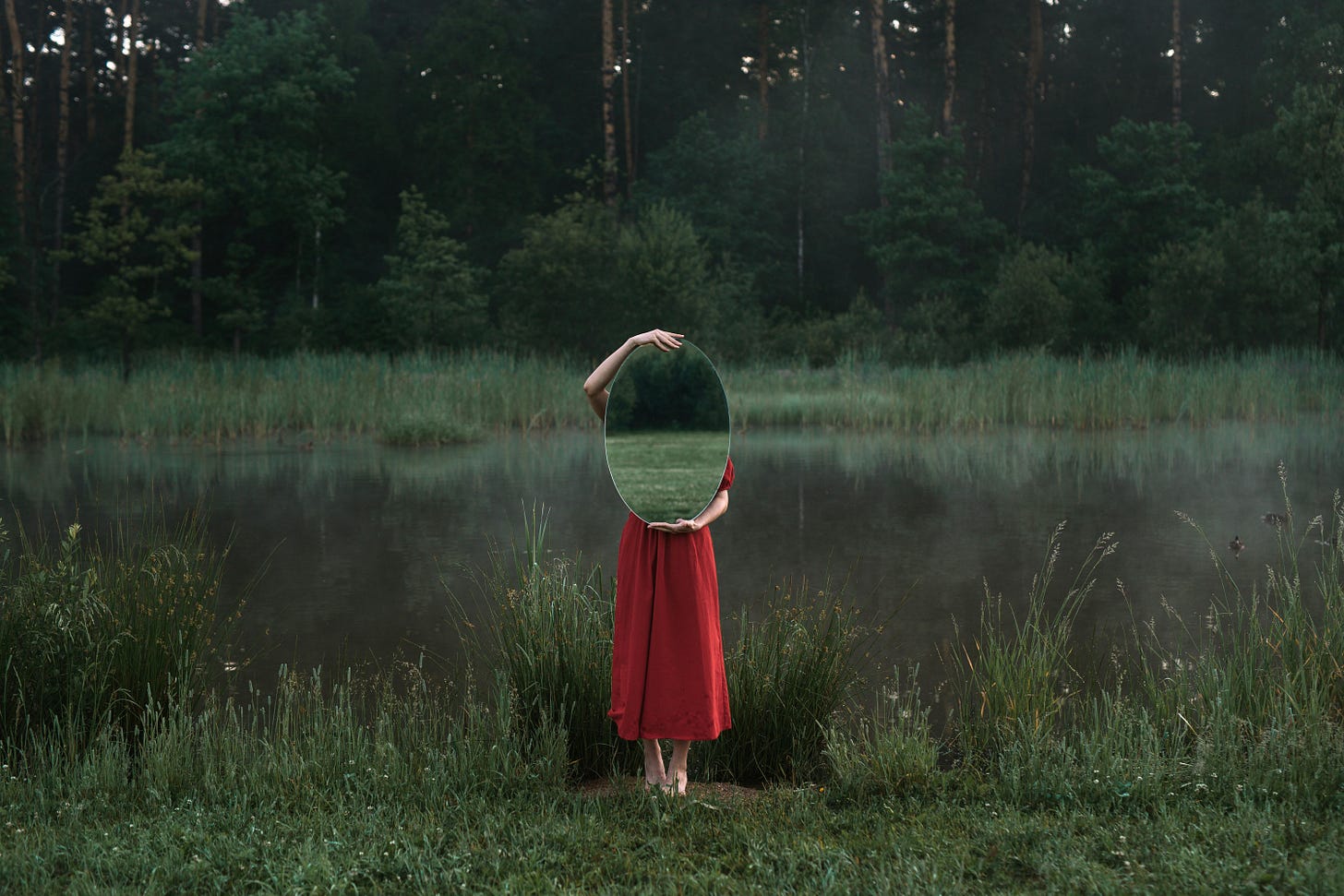 A woman standing in front of a pond holding a mirror in front of her that reflects the scenery around her.