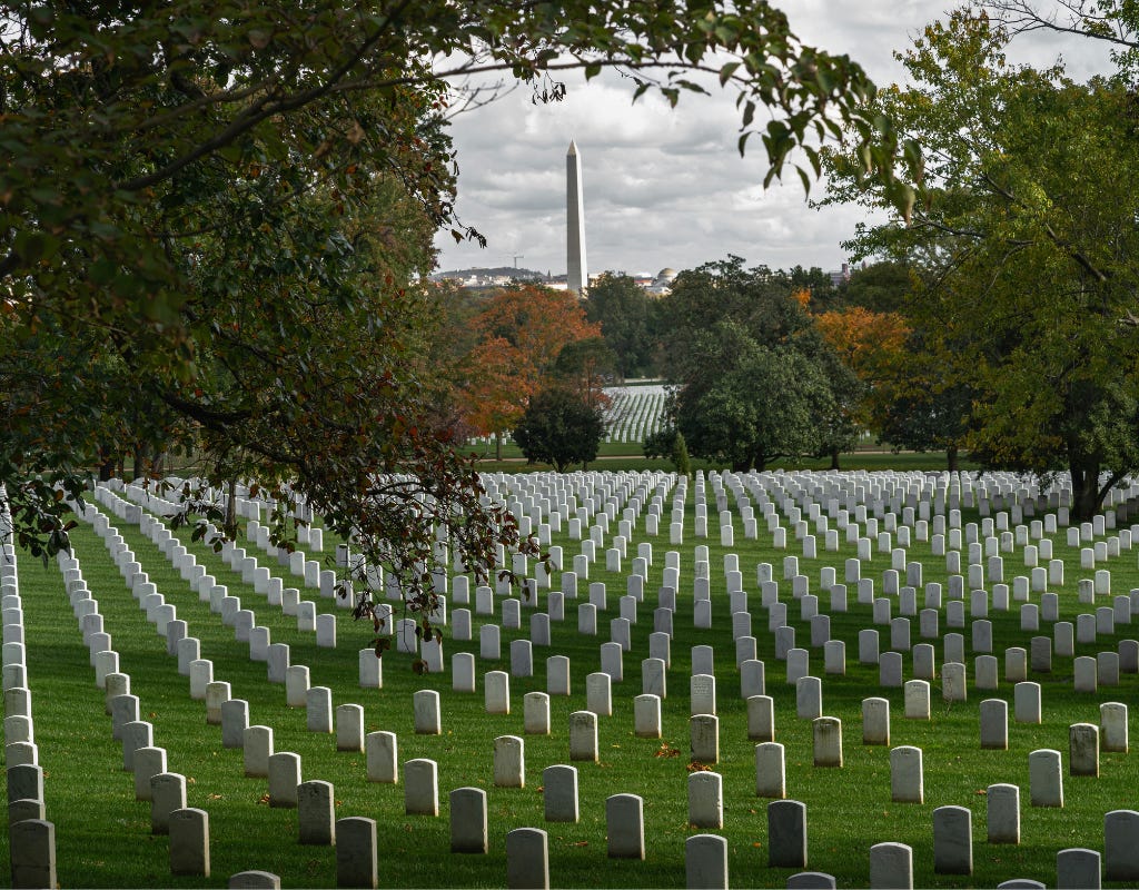 White gravestones on a hillside of Arlington National cemetery in the foreground surrounded by a mixture of green and orange leaved trees. Another field of white graves can be seen as the hill gently slopes downward. The Washington monument rises in he background under gray cloudy skies.