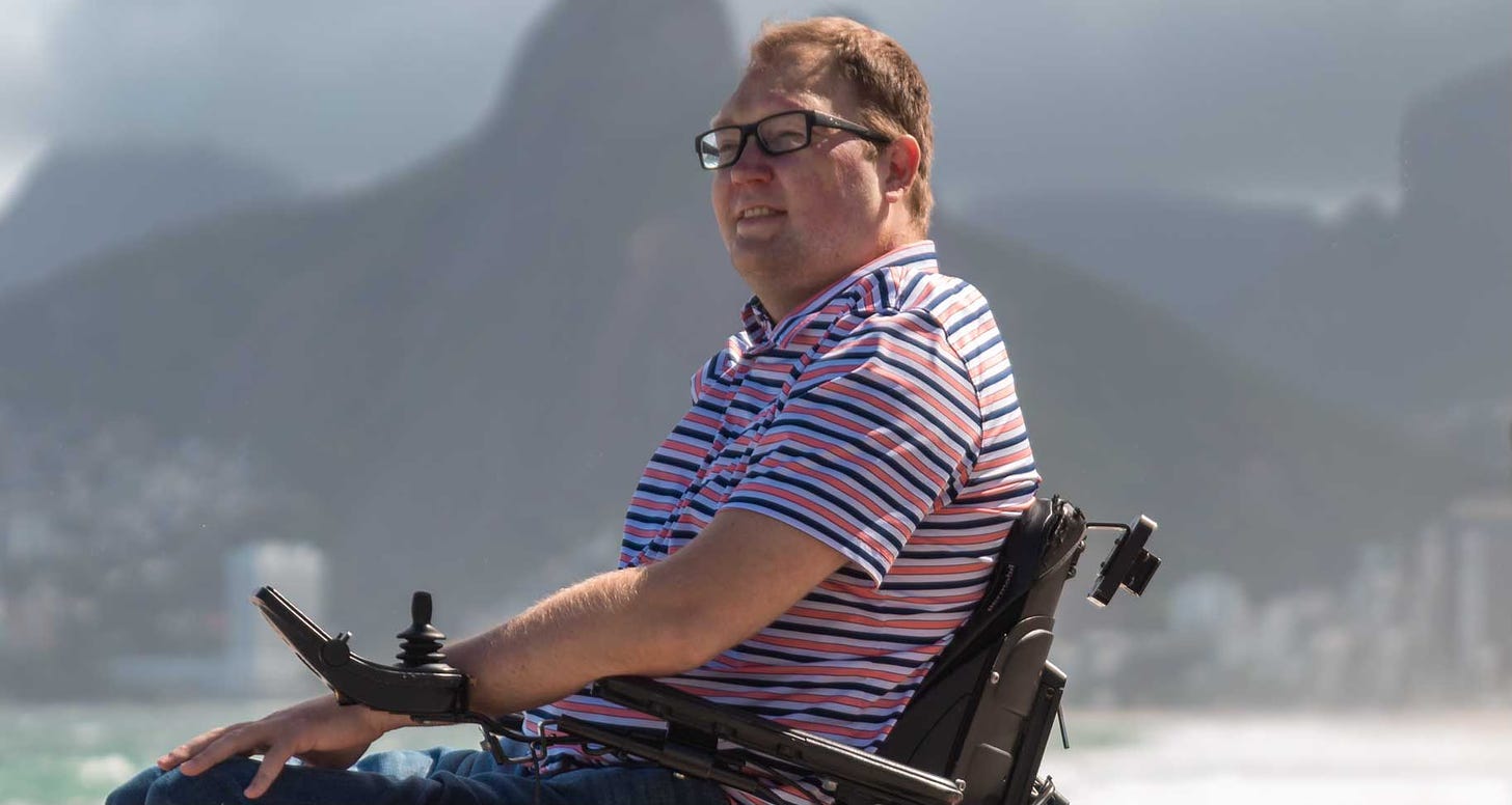 John seated in his wheelchair next to a beach with mountains in the background.