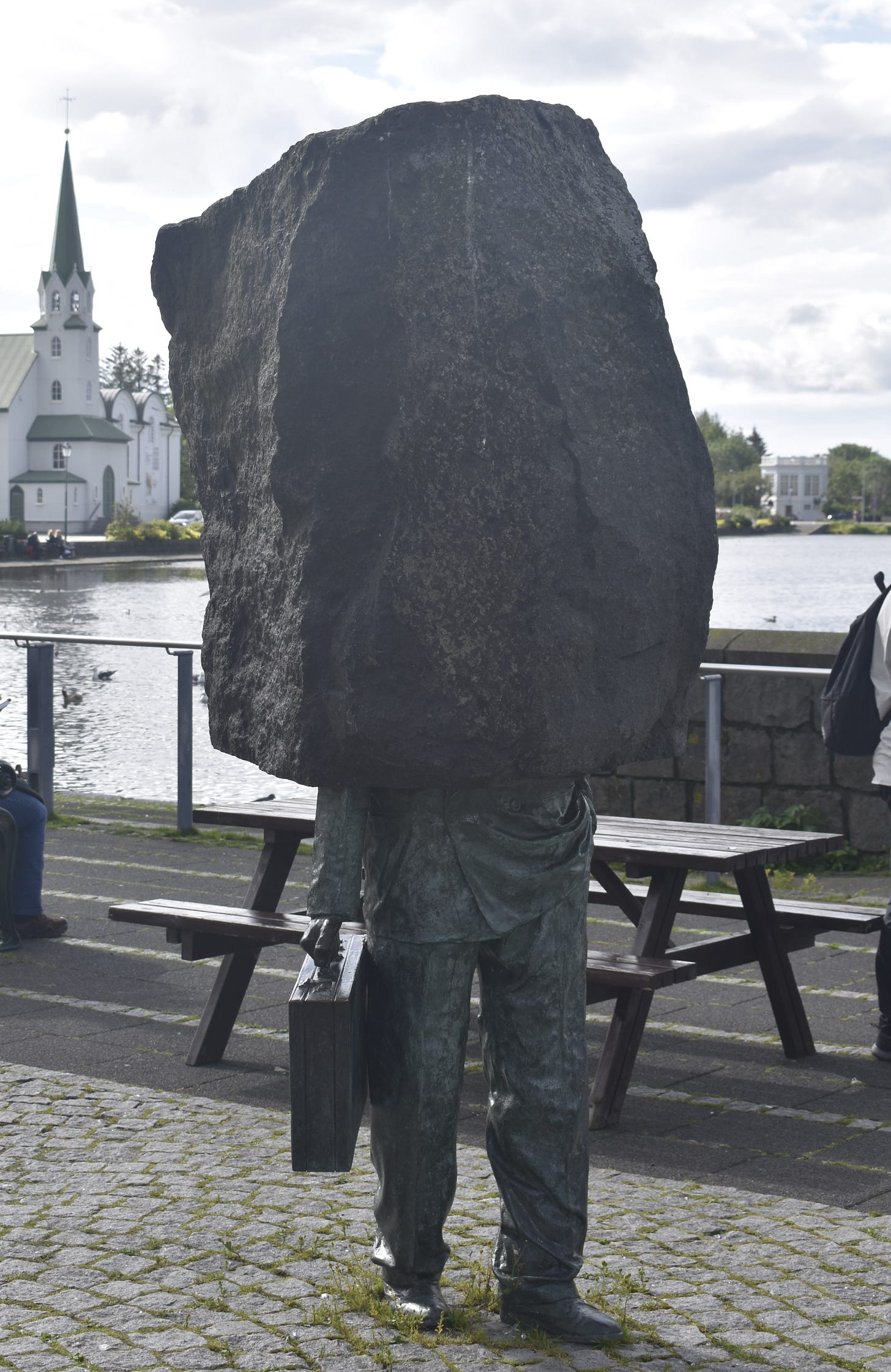 Statue of a lump of stone on top of the waist and legs of a man holding a briefcase. In the background is a lake and a white church.