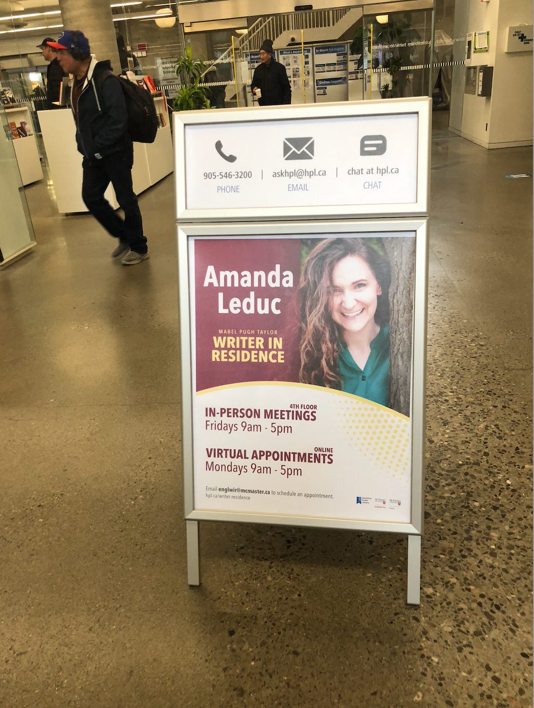 An A-frame sign sits in the middle of a busy library concourse. The sign advertises the photo of a smiling brown-haired white woman wearing a green shirt. Text on the sign says AMANDA LEDUC: MABEL PUGH TAYLOR WRITER-IN-RESIDENCE