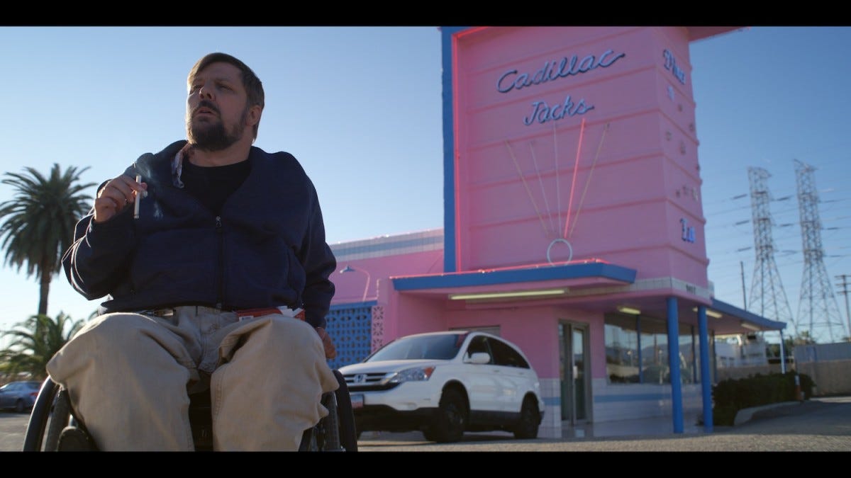A white disabled wheelchair using man is smoking a cigarette outside a pink motel, looking a bit forlorn.