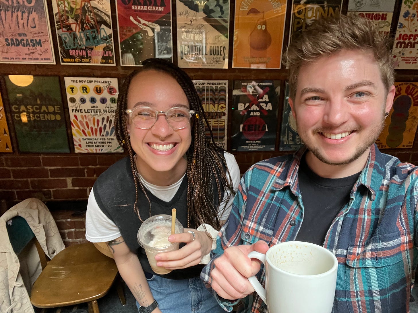 Oak and Jack pose with their coffee cups in front of a poster-laden wall