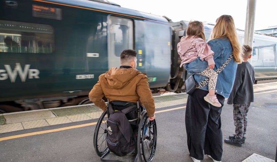 train platform.  GWR train in station.  A man in a wheelchair wearing a brown coat and short dark hair.  His hands are on the wheels ready to push.  A blonde haired woman wearing a denim jack and dark trousers is holding a young child with a pink top and leopard print leggins.  To her right is a boy with short hair, grey top and camo trousers.  