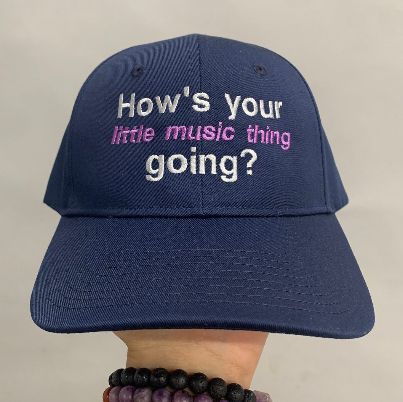 This image shows a navy blue baseball cap with an embroidered message on the front that reads, "How's your little music thing going?" The text is in white, with "little music thing" highlighted in pink. The phrase appears to carry a playful or perhaps slightly sarcastic tone, often reflecting how people might downplay or underestimate someone's creative endeavors, like a music project.