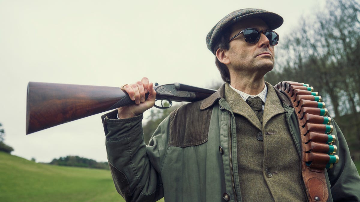 David Tennant in costume wearing hunting gear and flat cap, and carrying a rifle and ammunition over his shoulder