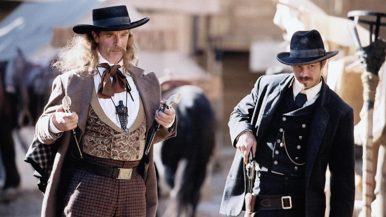 This image shows Wild Bill Hickok (played by Keith Carridine, left) and Seth Bullock (played by Timothy Olyphant, right) with their hands on their pistols while standing in Deadwood's main thoroughfare. Both men stare at something or someone off-camera.