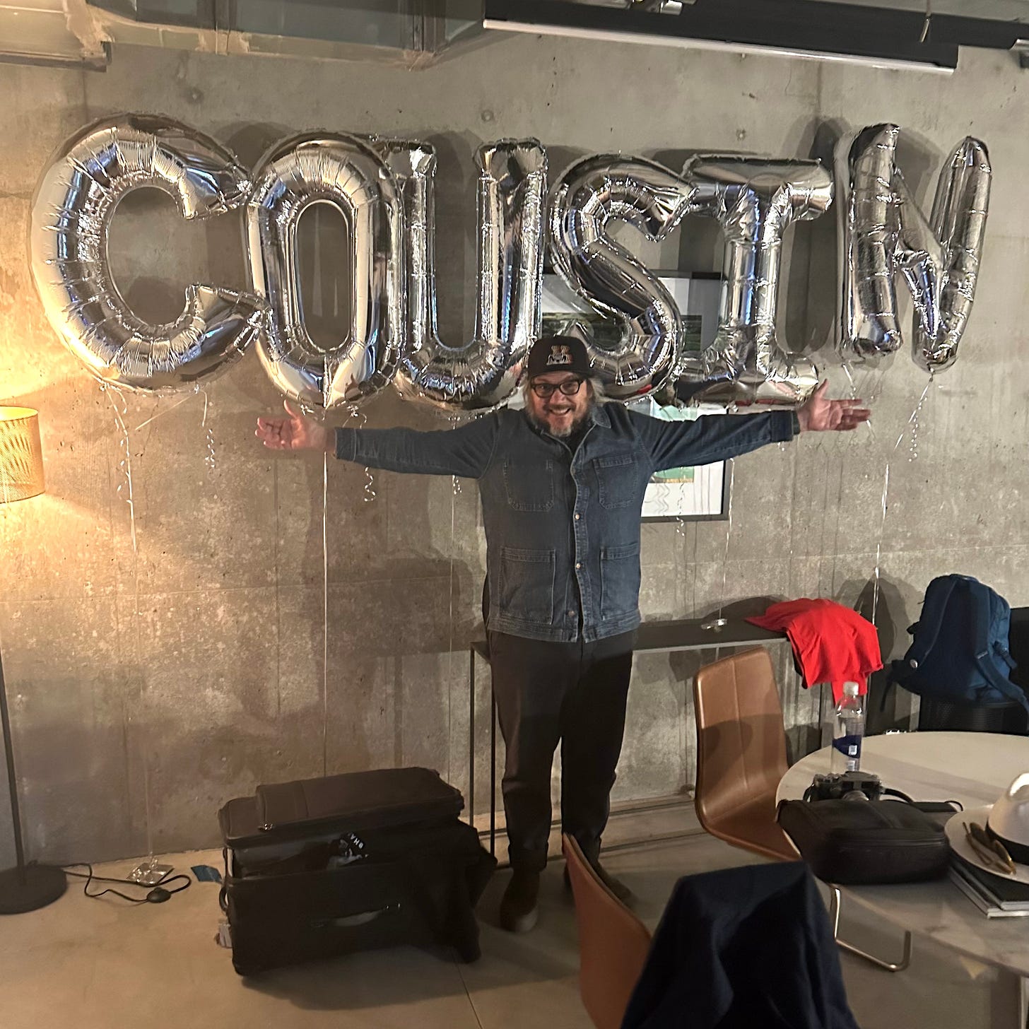 Jeff poses, arms outstretched, in front of silver balloons that spell COUSIN.