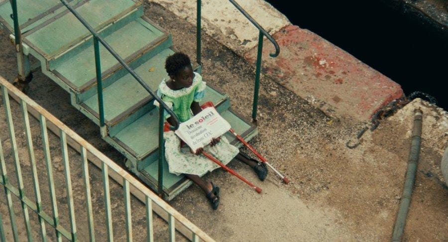 Sili, a young black disabled girl sits at the bottom of some outdoor steps. She’s wearing a beautiful green and blue dress, has her red crutches resting by her sides, and is holding a pile of Le Soleil newspapers.