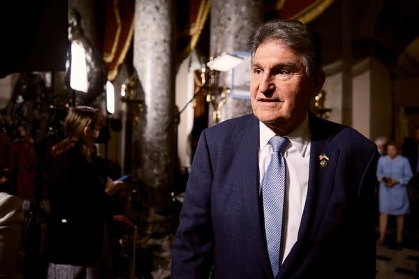 Joe Manchin leaving the House chamber. He is wearing a blue suit, white shirt and blue tie. Cameras with lights are set up behind him.