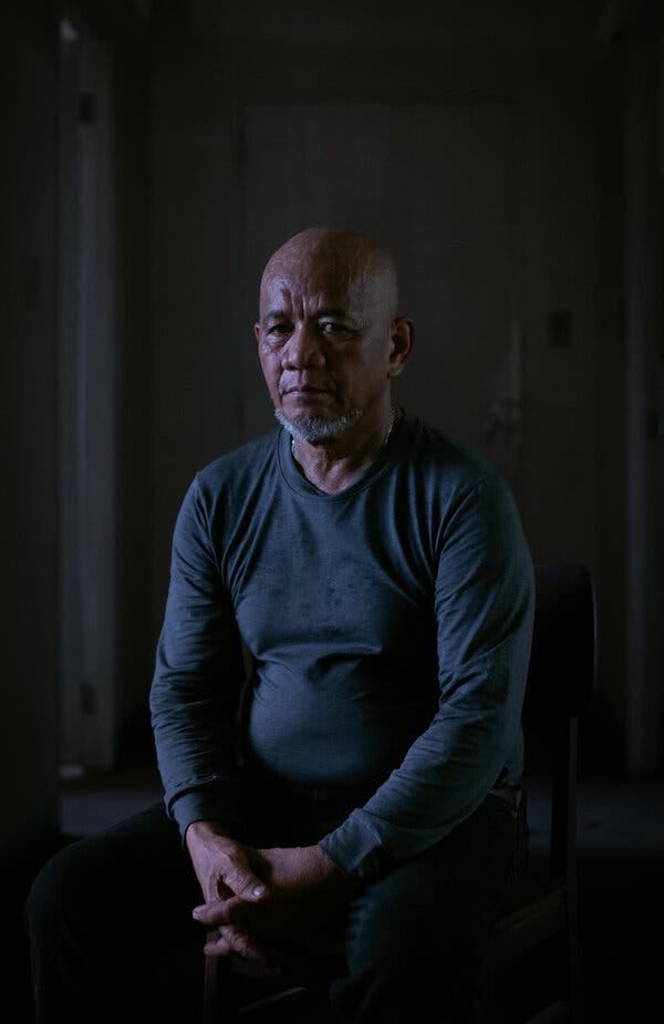 A frowning bald man sits in a darkened room with his hands folded.