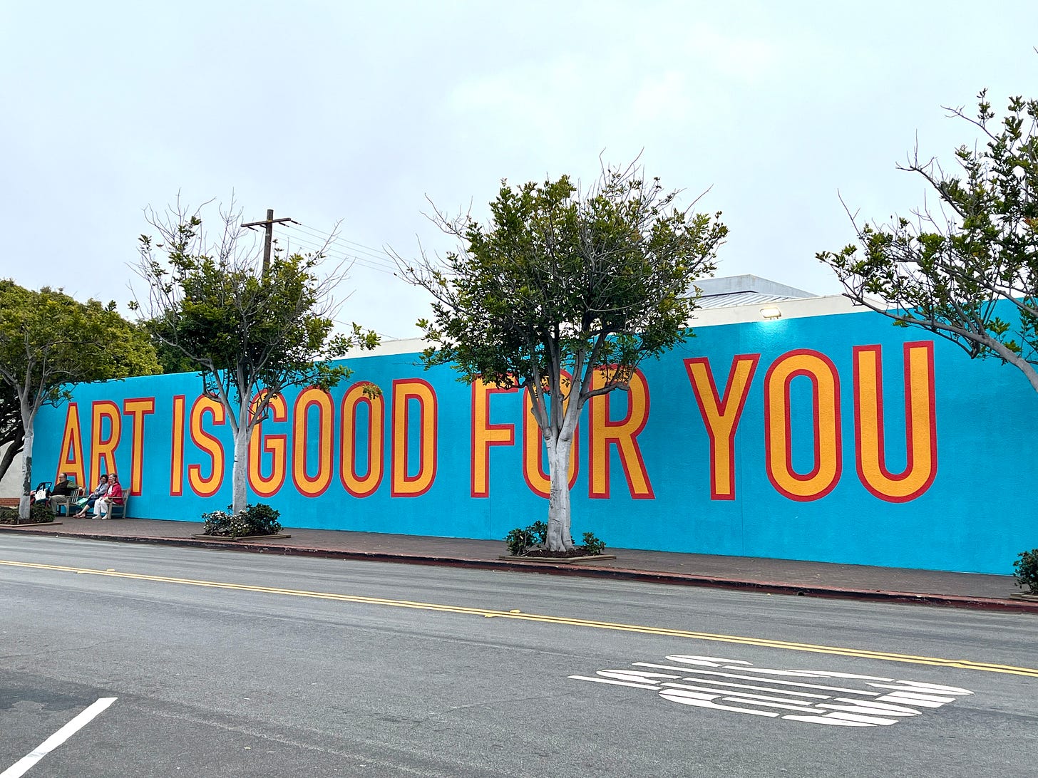 Long blue wall with word painted in bright orange and all caps: ART IS GOOD FOR YOU