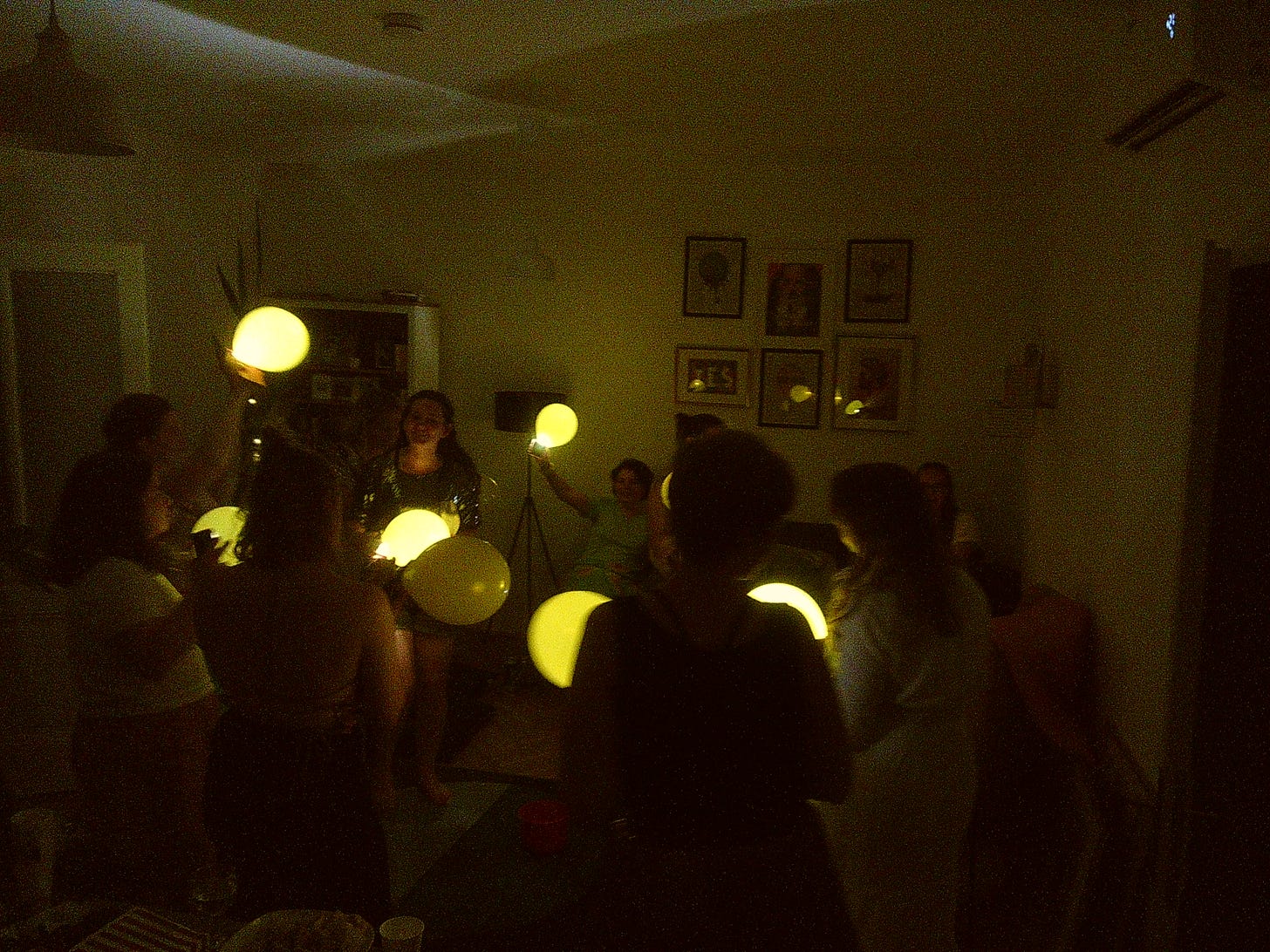 Grainy photo showing a group of women in an apartment with the lights switched off, each holding a yellow balloon with their phone torch shining through it to look like a glowing orb. 