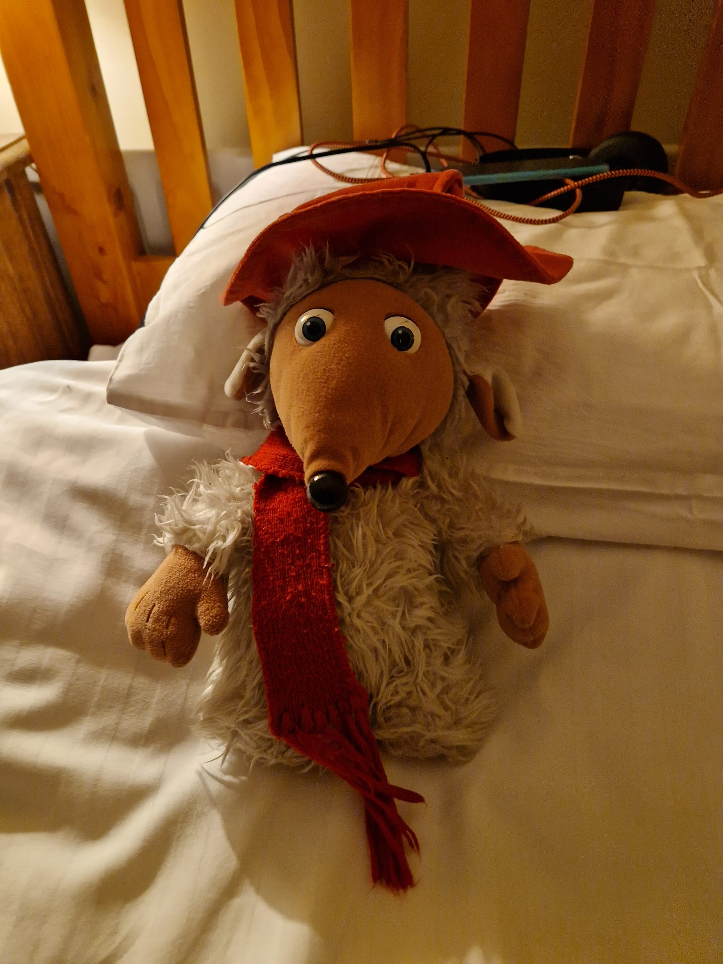My cuddly toy of Orinoco, the Womble character. He has blue eyes, has grey fur, an orange face and snout, and floppy ears. He wears a reddish brown hat and a red woollen scarf. He is perched on a single bed with white sheets. Behind him is an iPod with a large set of Sony headphones attached to an orange charge cable. 