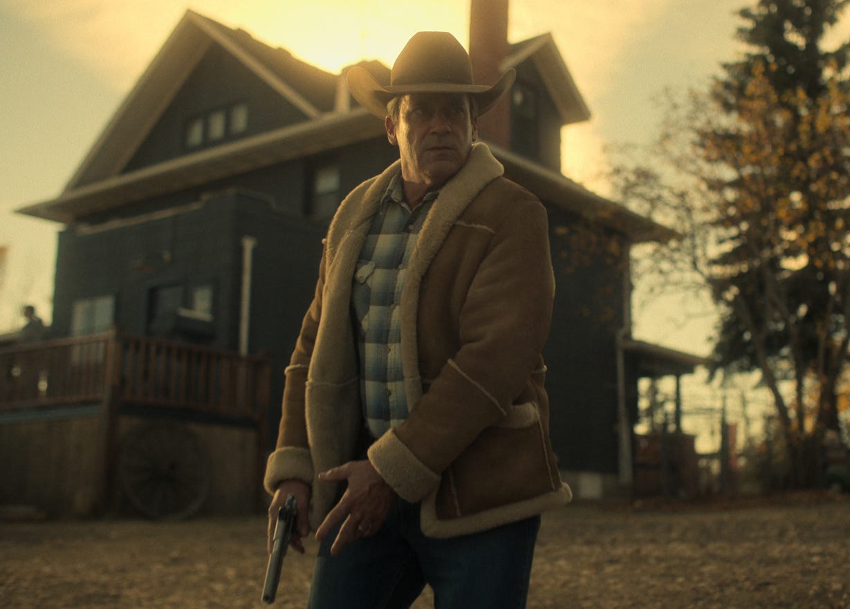 a man in a wide brim hat and brown leather jacket holds a gun in front of a house