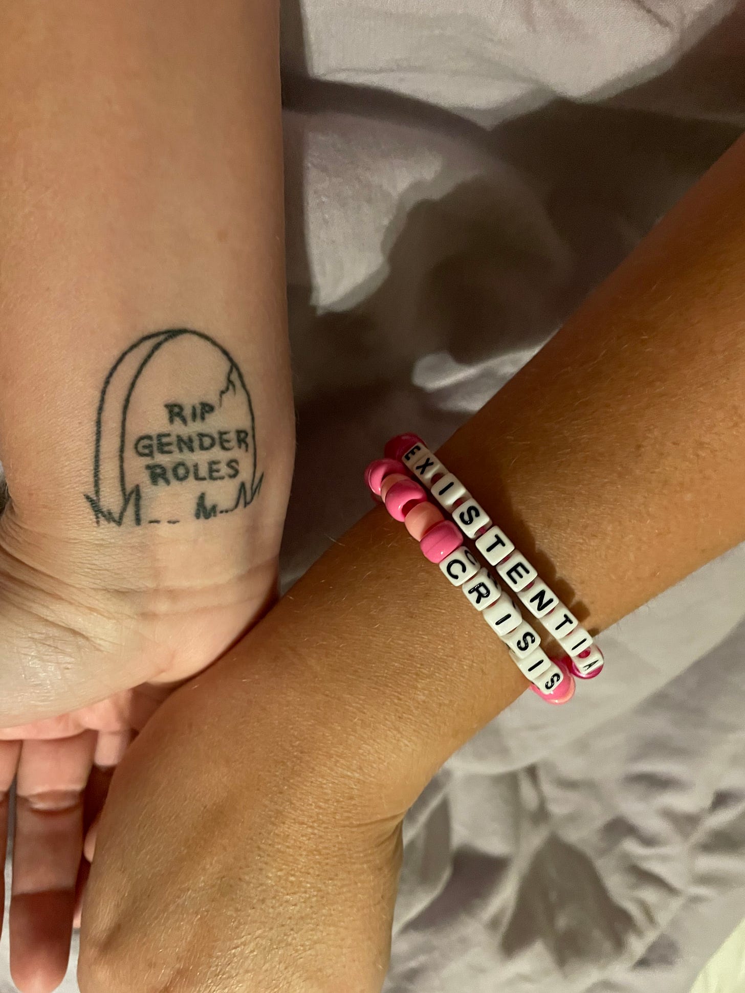 two white arms, one has a black and white tattoo of a tombstone that says RIP gender roles, the other arm has two pink beaded bracelets with white letter beads that say existential crisis on them