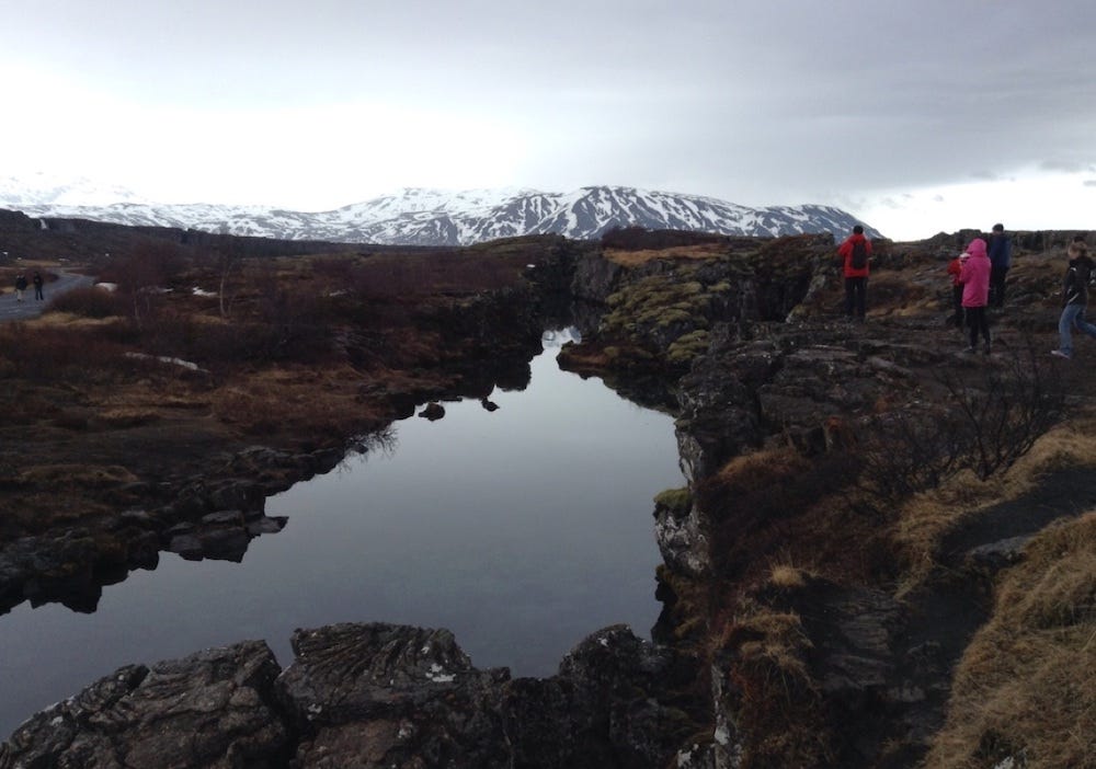 A stream runs through the arid landscape. Snow-capped mountains behind.