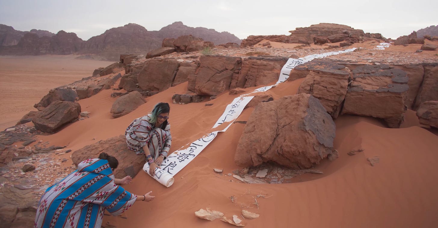 Adelle unrolls a scroll with Arabic and Chinese script on a desert hill