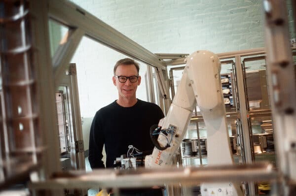 Steve Ells in black-rimmed glasses and a long-sleeved black T-shirt stands behind a large white robotic arm.