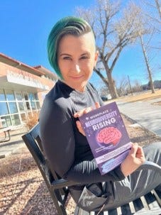 Lyric, a pale skinned multiracial, nonbinary human, with short green hair and shaved sides is sitting on a bench in front of a brick building that says "school of business" - in Lyric's hand is a purple book with a pink brain on it that says Workplace NeuroDiversity Rising