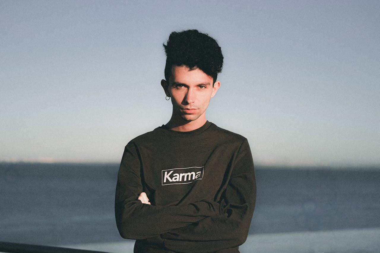 A man wearing a black t-shirt with the word "karma" on it stands in front the backdrop of sea and sky. A man wearing a black t-shirt with the word "karma" on it stands in front the backdrop of sea and sky.
