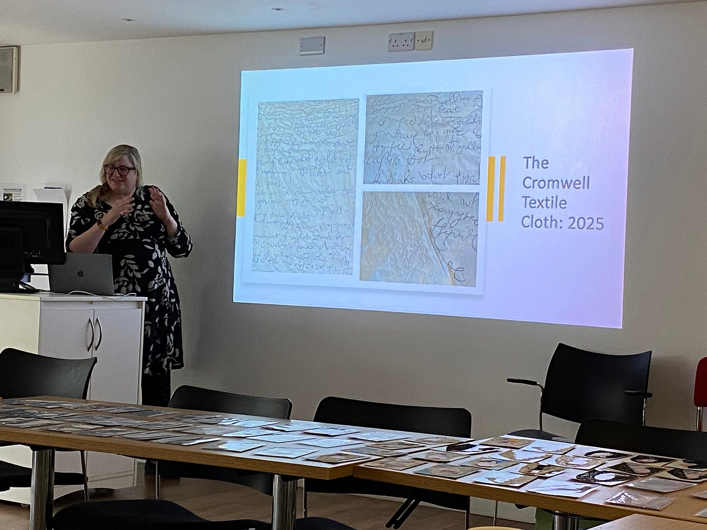 A white woman stands in front of a projection of The Cromwell Textile Cloth 2025 - a stitched piece in cream and black showing 16th century text.