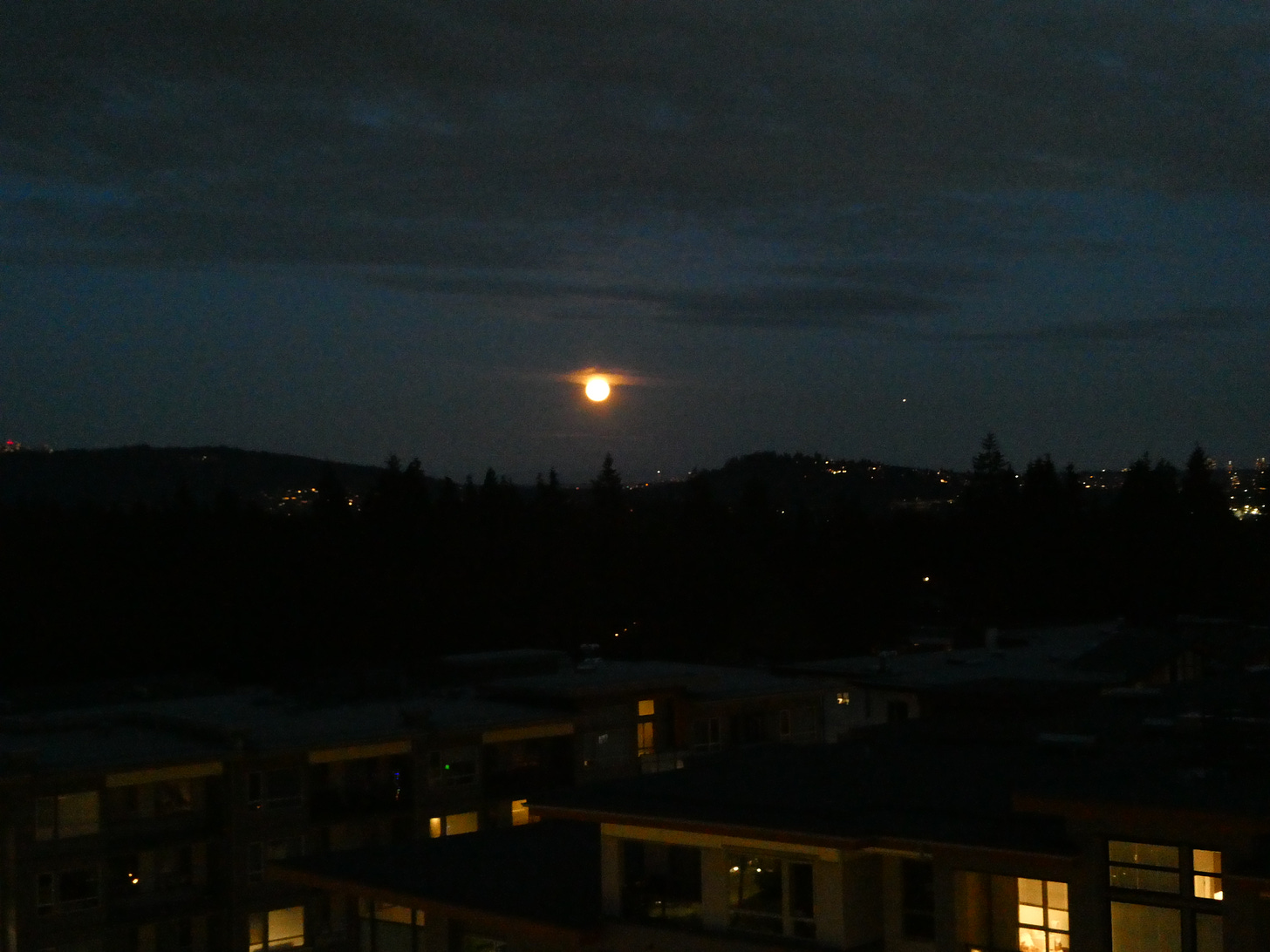 Low and orange moon in dark sky over low buildings.