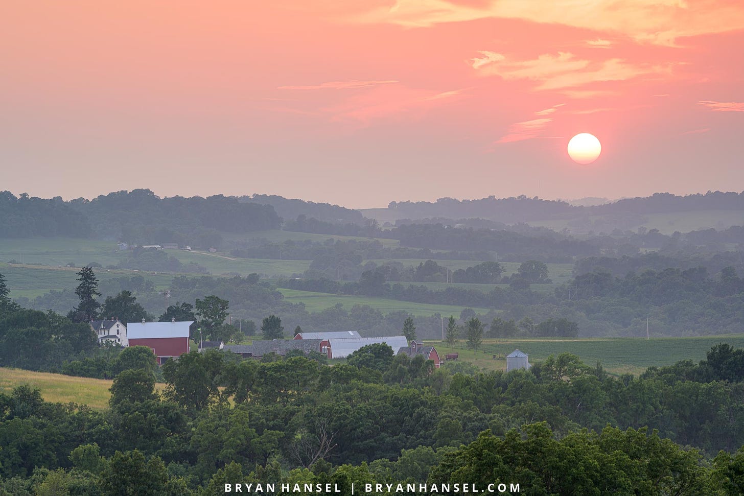 A picture of sunset over a patchwork of trees and farm fields. The orange sundisc is in the sky.