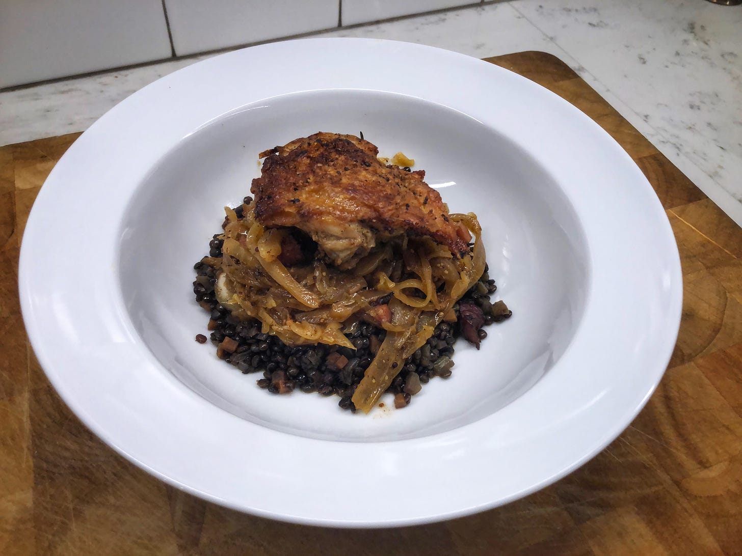 On a kitchen counter, a bowl with a chicken thigh with crisp skin sits atop some braised cabbage and bacon, which sits atop a bed of beluga lentils