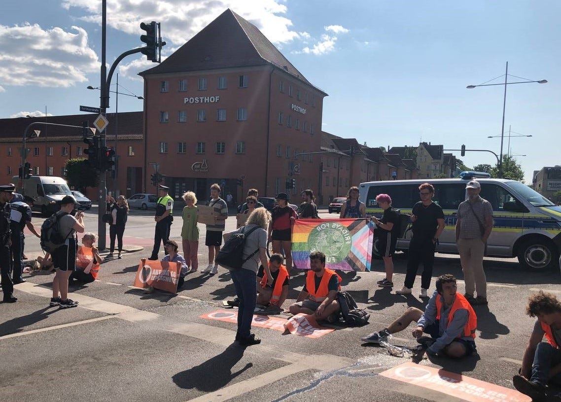 Bild 1: Unterstützer*innen stehen hinter und neben der Straßenblockade. Manche halten Schilder oder Flaggen von anderen Gruppierungen wie Fridays for Future.