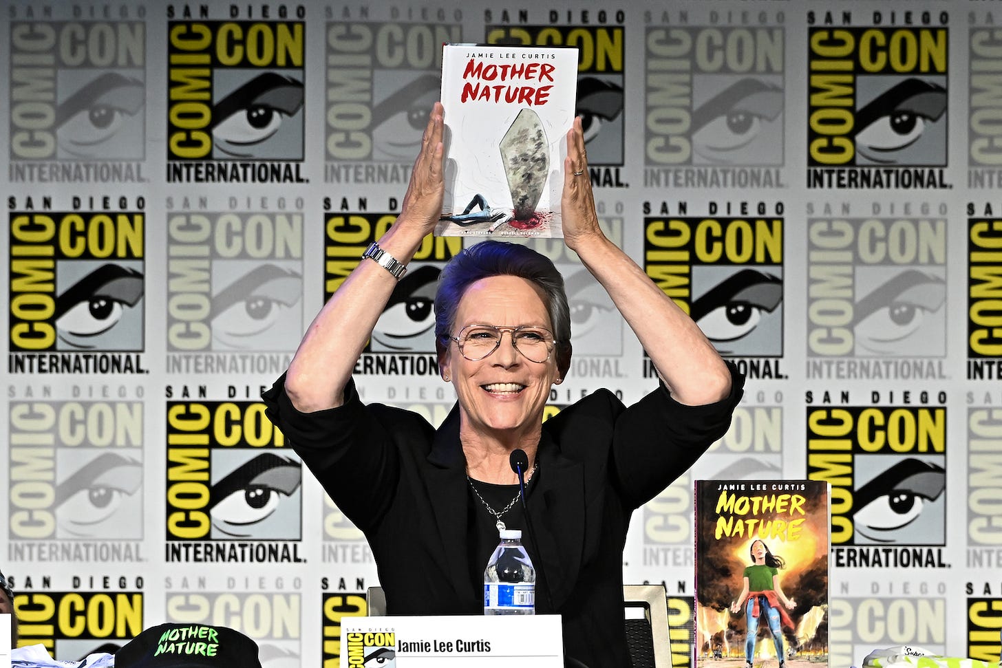 Actor Jamie Lee Curtis holds a hardcover comic book above her head called Mother Nature. She is wearing a black shirt and glasses. She sits at a table; behind her is the Comic Con logo