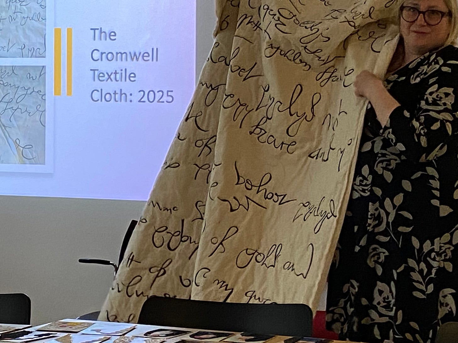 A white woman holds up a piece of large textile art, but is unsuccessful in holding it straight and fully visible.