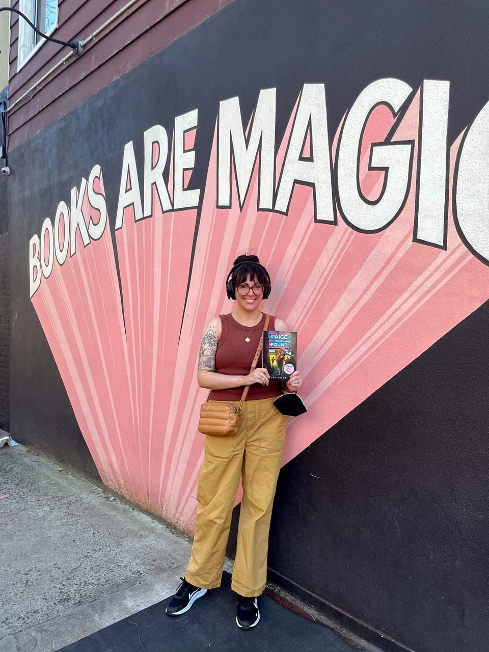 Jen Wilde, a white nonbinary femme, stands outside of Books Are Magic in Brooklyn, holding a copy of her book, Paige Not Found.