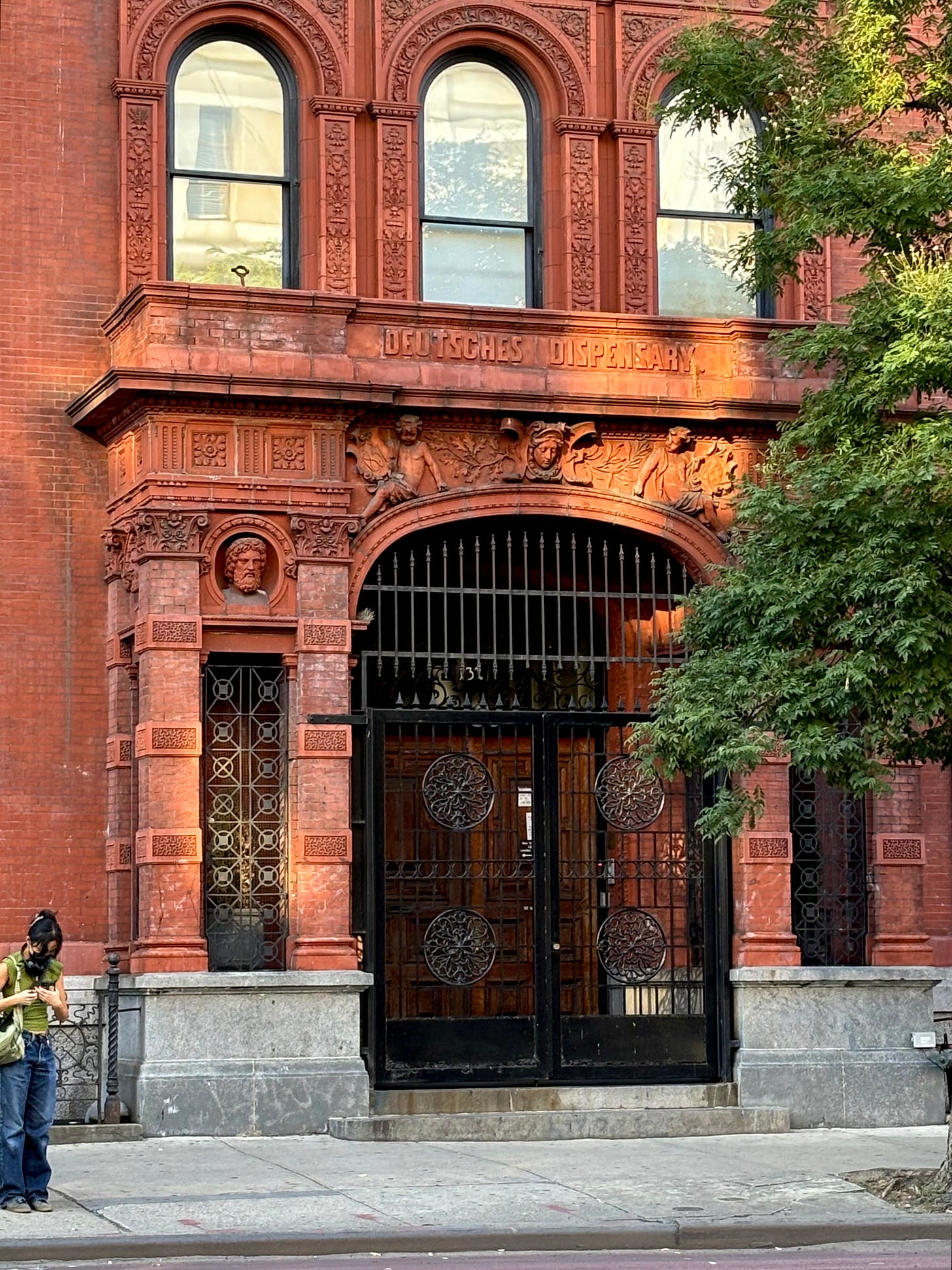 The engraved entrance to the dispensary. It is marked DEUTSCHES DISPENSARY over winged-cherubs are over a curved gate. There are three arched windows above and a blooming tree to the side.