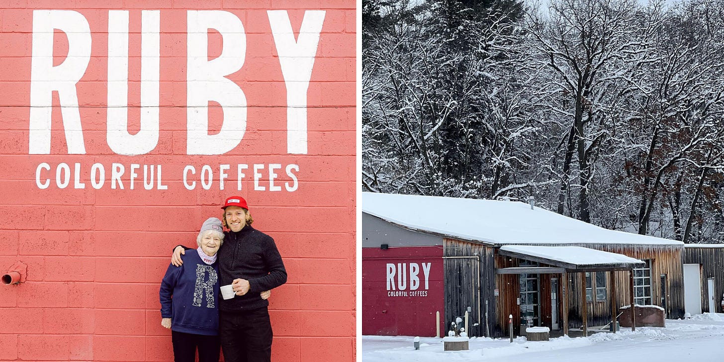 A young man holding a coffee mug has his arm around his grandmother as they lean up against a red concrete block wall. Right - A worn wood sided building with a low-slung roof and side walls painted a deep red is covered in a layer of bright white snow. Leafless snow-covered trees fill the background.