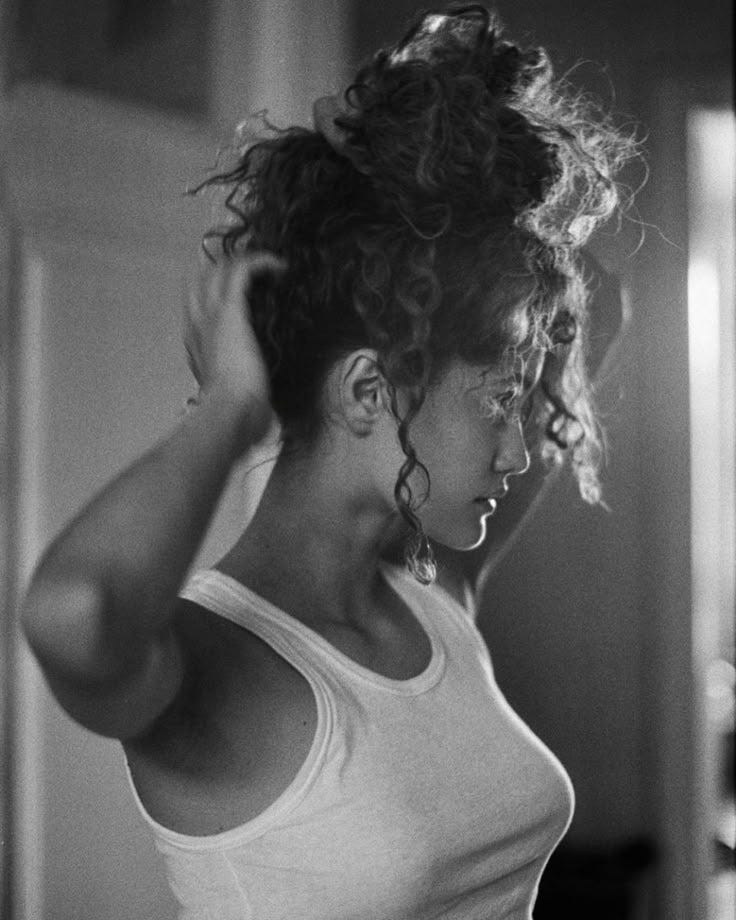 This may contain: a black and white photo of a woman with curly hair in a tank top looking at her cell phone