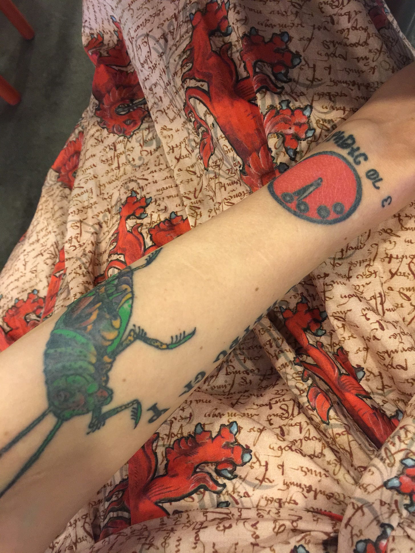 close up of a white person's wrist showing a red doomsday clock tattoo where a watch would typically sit. Below it is text reading 3 minutes to midnight in deseret alphabet. another tattoo of a grasshopper is just above it on the forearm.