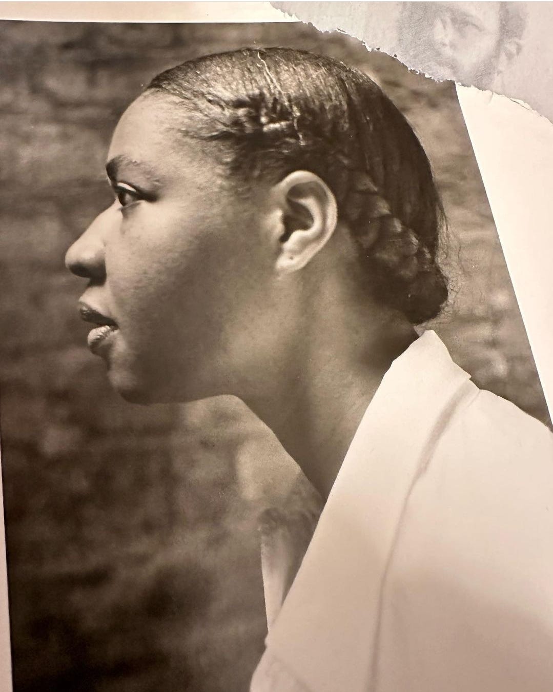 A black and white photo of a younger Jamaica Kincaid in profile. Her hair is straightened in a side braid, wearing a white blouse.