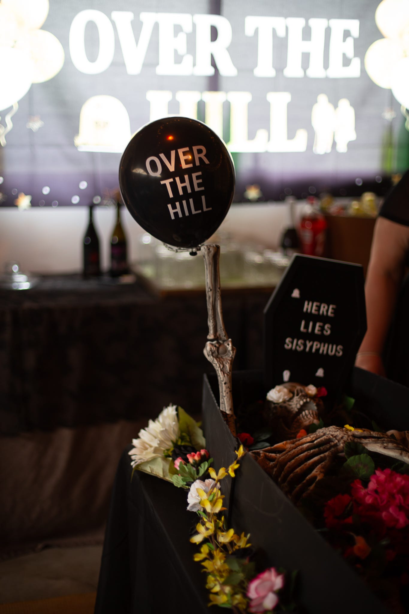 in the background is a banner that says over the hill. there is a fake coffin with a fake skeleton in the middle of the room, the fake skeleton hand is holding a black balloon with white letters that say over the hill