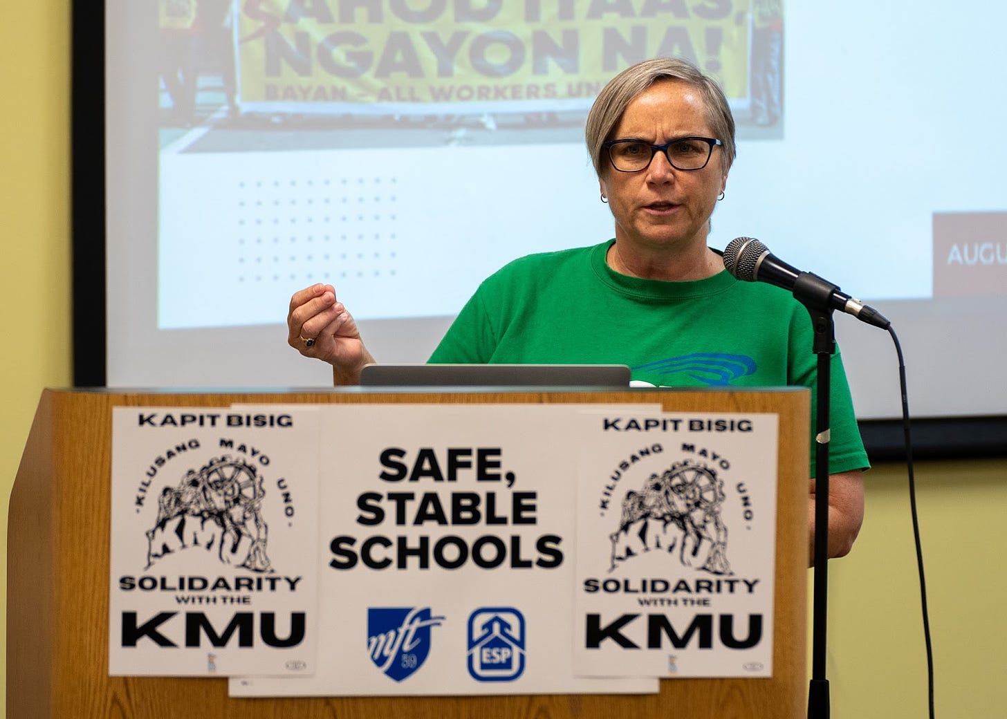 A white woman with short gray hair and glasses wearing a green tee shirt stands behind a wooden podium with posters reading "Kapit Bisig solidarity with the KMU" and "Safe, stable schools"