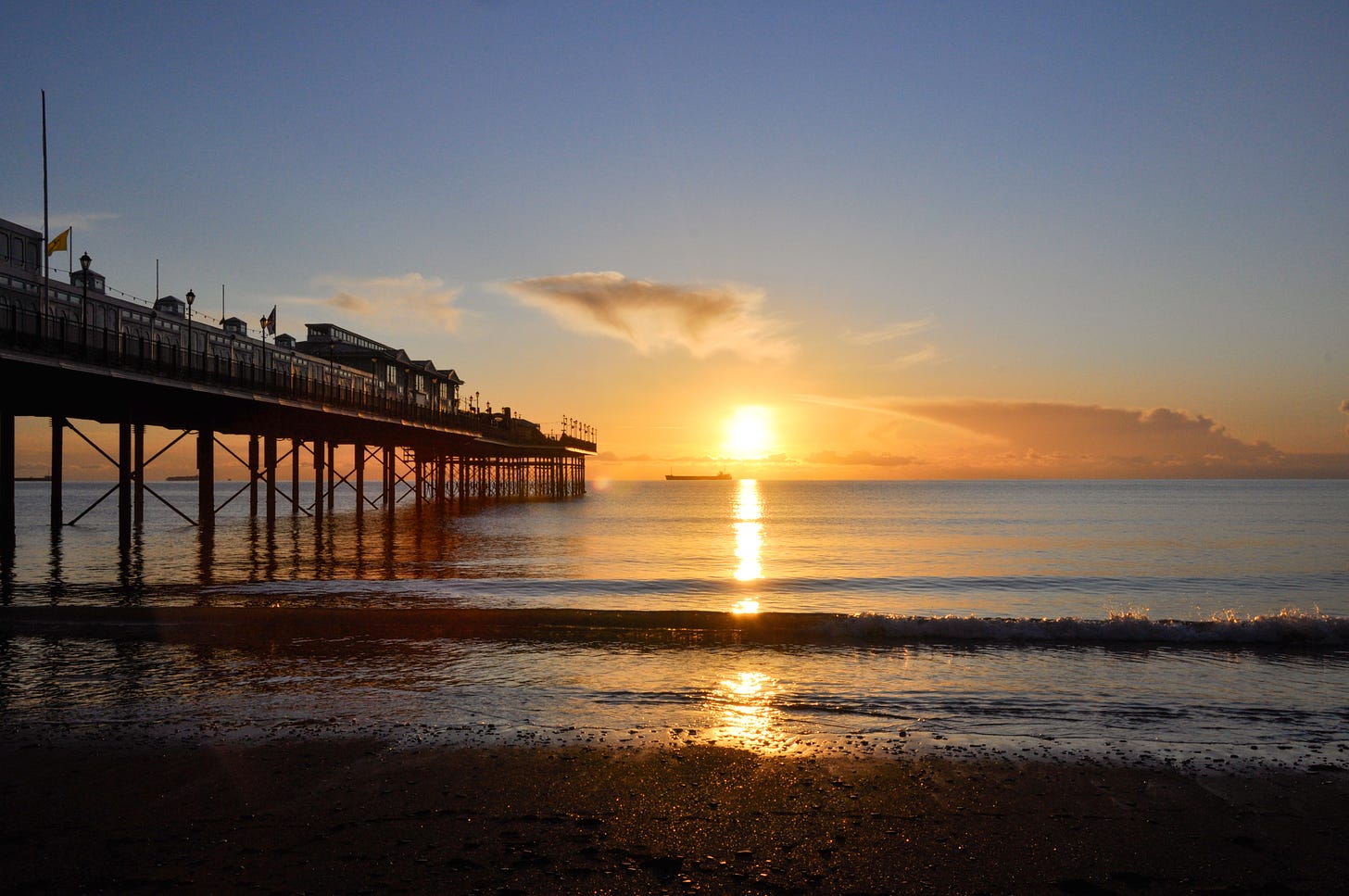 An image of the sunrise above the sea. Paignton pier is to the left, stretching out toward the sun. The sky is orange close to the horizon and clear blue further out. The sea reflects a column of warm yellow light.