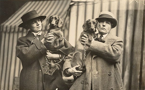 Black and white photograph of Una Troubridge and Radclyffe Hall holding their dogs in 1923. Both women are dressed in woollen overcoats and wide-brimmed hats. Black and white photograph of Una Troubridge and Radclyffe Hall holding their dogs in 1923. Both women are dressed in woollen overcoats and wide-brimmed hats.