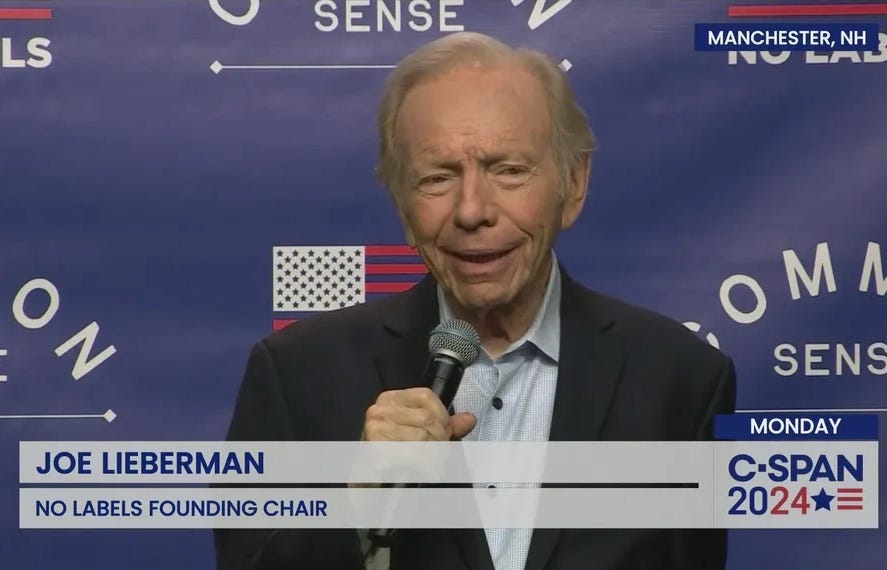 CSPAN screenshot of Joe Lieberman being interviewed about No Labels, in Concord, New Hampshire. He holds a microphone and stands in front of a background printed with flags and the slogan 'Common Sense'