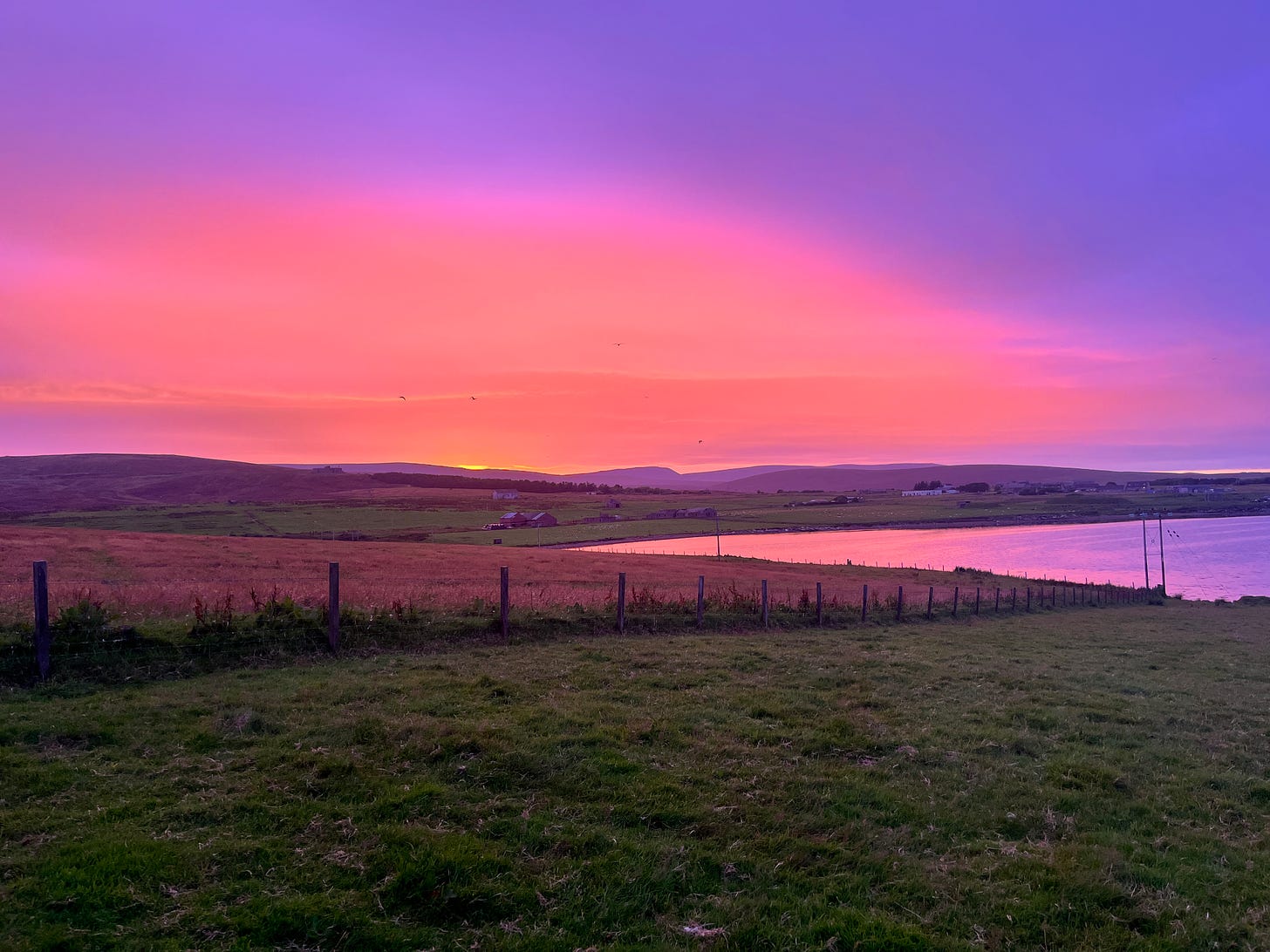 Sunset looking across grass, over a small inlet & onto hilss in the background. Colours of purple, pink & orange