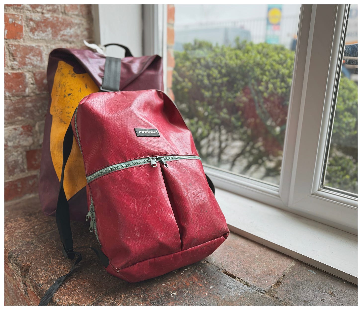 A colorful FREITAG backpack leans against a brick wall next to a window overlooking some bushes. 