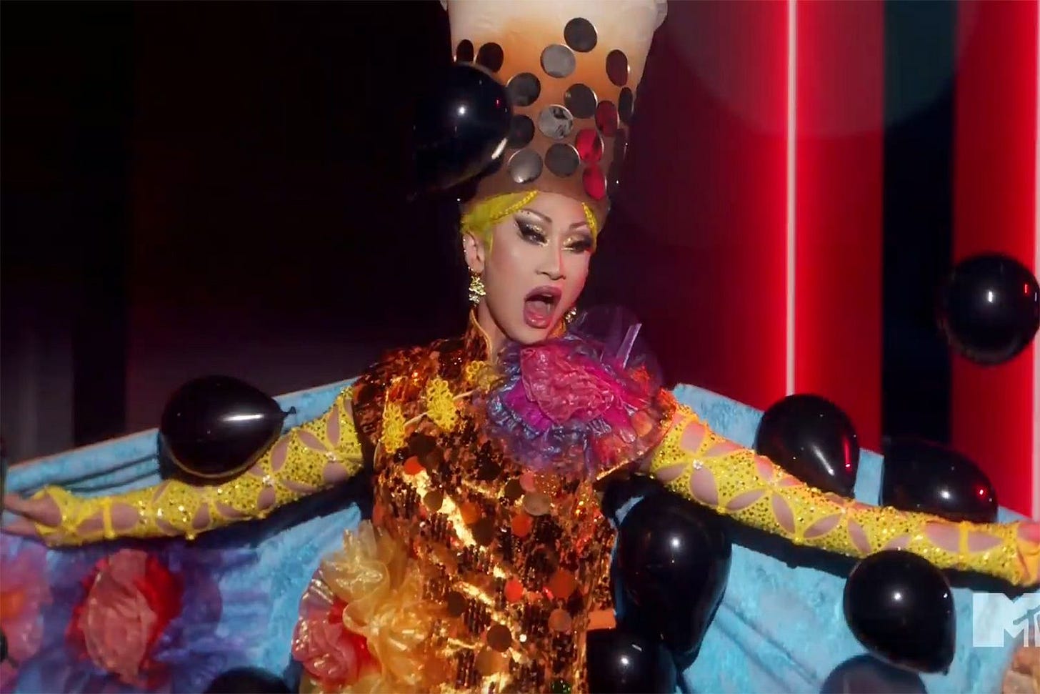 A drag queen performs with in a milk tea-shaped headpiece, a shimmering gold dress, and a blue-lined cape that opens to release black balloons into the air A drag queen performs with in a milk tea-shaped headpiece, a shimmering gold dress, and a blue-lined cape that opens to release black balloons into the air